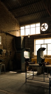 An industrial workshop with brick walls and a high ceiling. The space is lit by natural light coming through large windows, highlighting metal ventilation pipes and workbenches. Some tools and machinery are visible and organized around the room.