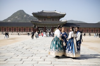 A group of people wearing traditional hanboks are gathered in the foreground while others walk along a wide stone path leading to an ornate, traditional Korean palace. Mountains rise in the background under a clear blue sky.