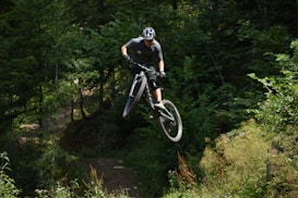 A mountain biker is caught in mid-air while performing a jump in a forest setting. The scene is filled with lush green trees and undergrowth, creating a sense of adventure and motion.