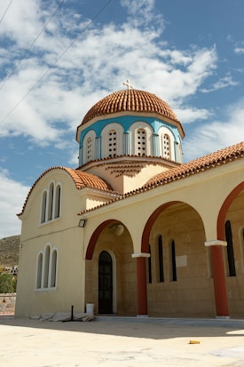A traditional church with arched windows and a colorful domed roof adorned with a cross. The building features cream-colored walls and red-orange roofing tiles, creating a striking contrast against a bright blue sky with scattered clouds.