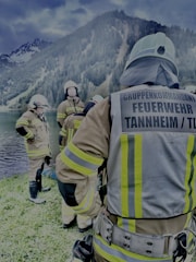 Several firefighters in full gear are gathered near a lakeside with forested mountains in the background. They appear to be preparing or finishing a task, with one firefighter facing away from the camera and the others engaged in conversation.