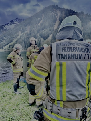 Several firefighters in full gear are gathered near a lakeside with forested mountains in the background. They appear to be preparing or finishing a task, with one firefighter facing away from the camera and the others engaged in conversation.