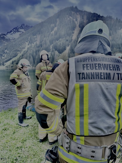 Several firefighters in full gear are gathered near a lakeside with forested mountains in the background. They appear to be preparing or finishing a task, with one firefighter facing away from the camera and the others engaged in conversation.