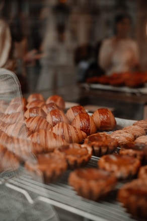 A display of freshly baked pastries, including croissants and other baked goods, behind a window. There is a warm, inviting atmosphere with soft lighting and reflections visible on the glass.
