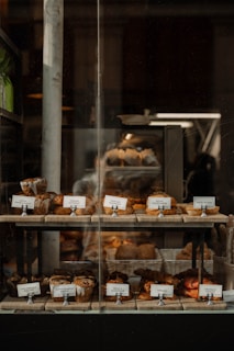 A bakery display window featuring an assortment of pastries and bread. The shelves are lined with various baked goods like croissants, muffins, and loaves, each accompanied by small name cards. The display is warmly lit, creating a cozy and inviting atmosphere.