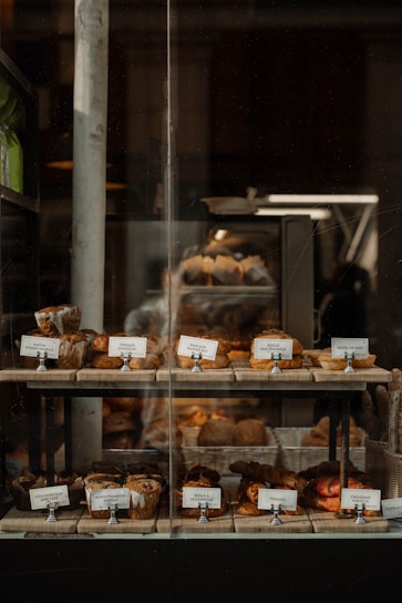 A bakery display window featuring an assortment of pastries and bread. The shelves are lined with various baked goods like croissants, muffins, and loaves, each accompanied by small name cards. The display is warmly lit, creating a cozy and inviting atmosphere.