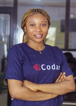 A person with braided hair is standing with their arms crossed, wearing a navy blue shirt with the word 'Codar' printed on it. The background appears to be an indoor setting, possibly an office.