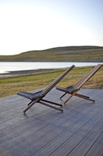 Two wooden deck chairs are positioned on a wooden patio overlooking a serene body of water with gentle hills in the background. The setting appears peaceful, likely capturing a moment near sunset with soft lighting on the landscape.