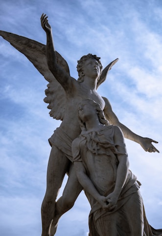 A detailed sculpture depicting an angelic figure with wings and outstretched arms, accompanied by a crouching person. The angel gazes upwards, while the person at their feet appears pensive, holding a rosary. The backdrop features a partly cloudy blue sky.