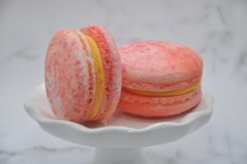 Two pink macarons with speckled patterns sit on a white, scalloped-edge cake stand. A yellow filling is visible between the almond cookies, giving a vibrant contrast to the pastel hues. The background is softly blurred, enhancing the focus on the macarons.