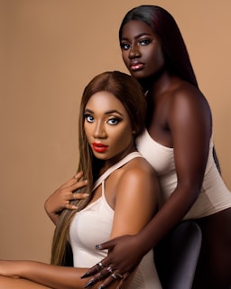 Two women are posing closely together against a neutral background. Both have long hair and are wearing light-colored tops. One is sitting while the other stands behind her, resting her arms on the seated woman's shoulder. They have bold makeup, with one featuring bright red lipstick.