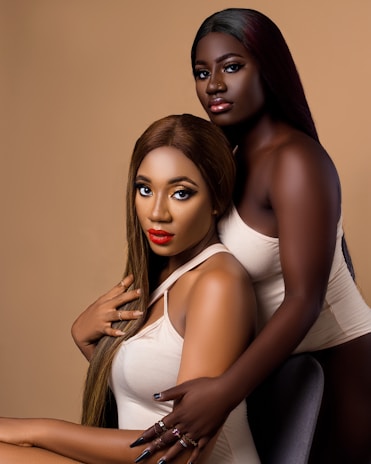Two women are posing closely together against a neutral background. Both have long hair and are wearing light-colored tops. One is sitting while the other stands behind her, resting her arms on the seated woman's shoulder. They have bold makeup, with one featuring bright red lipstick.