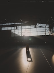 A dimly lit, large indoor space with a vast window grid allowing sunlight to cast sharp shadows on the floor. At the center, a prominent caution sign stands, illuminated by a stream of light.