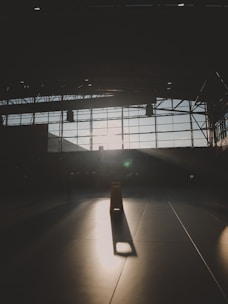 A dimly lit, large indoor space with a vast window grid allowing sunlight to cast sharp shadows on the floor. At the center, a prominent caution sign stands, illuminated by a stream of light.