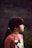 A young girl with dark hair tied in a ponytail is seen in profile view against a blurry outdoor background. She is wearing a reddish-brown shirt featuring a graphic of a cat with a bow.