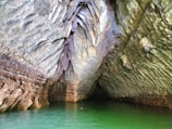 The image captures the interior of a cave with intricate rock formations. The ceiling and walls exhibit a variety of textures, appearing layered and rugged, with colors ranging from earthy browns to muted greens. The cave opens up to a pool of vibrant green water that reflects the surrounding rock structures.