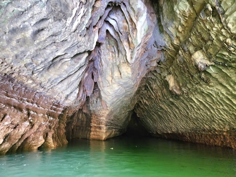 The image captures the interior of a cave with intricate rock formations. The ceiling and walls exhibit a variety of textures, appearing layered and rugged, with colors ranging from earthy browns to muted greens. The cave opens up to a pool of vibrant green water that reflects the surrounding rock structures.