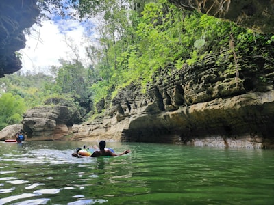 A lush green gorge with steep rock walls on either side, partially submerged in clear water. Two people, each on an inflatable tube, are relaxing and floating in the water. Vegetation is dense with a variety of plants and trees growing from the rocky surfaces. The sky is partly cloudy, allowing sunlight to peek through and reflect off the water's surface.