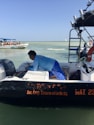 A person wearing a blue shirt is working on a small black and white boat on the water, with the text 'Soc. Coop. Ensueño del Caribe SCL' visible. In the background, another boat with several seated people wearing life vests is seen. The sky is clear and the water is calm.