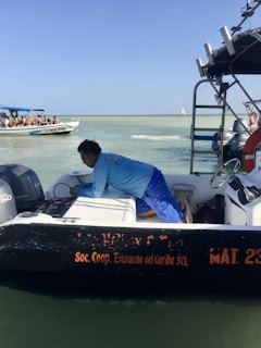 A person wearing a blue shirt is working on a small black and white boat on the water, with the text 'Soc. Coop. Ensueño del Caribe SCL' visible. In the background, another boat with several seated people wearing life vests is seen. The sky is clear and the water is calm.