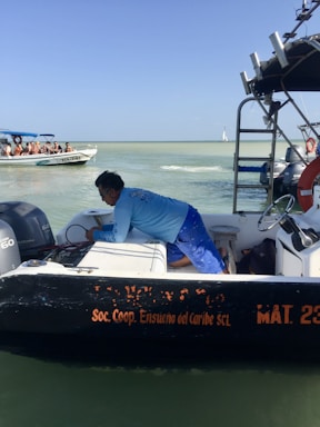 A person wearing a blue shirt is working on a small black and white boat on the water, with the text 'Soc. Coop. Ensueño del Caribe SCL' visible. In the background, another boat with several seated people wearing life vests is seen. The sky is clear and the water is calm.