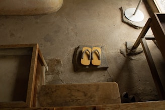 A pair of traditional wooden sandals, known as geta, are placed on a stone slab against a textured concrete floor. The setting appears to be indoors with wooden furniture framing part of the scene.