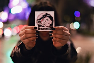 A person holds up a small square sticker in both hands against a blurred city backdrop with colorful bokeh lights. The sticker features an illustration of a dog wearing a space helmet with the word 'KASBAHN' printed beneath.