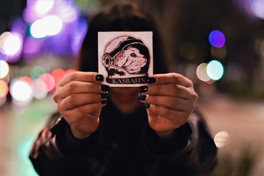 A person holds up a small square sticker in both hands against a blurred city backdrop with colorful bokeh lights. The sticker features an illustration of a dog wearing a space helmet with the word 'KASBAHN' printed beneath.