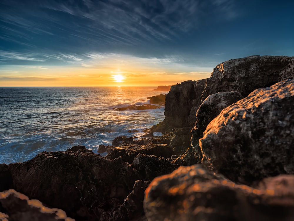 the sun is setting over the ocean on the rocky shore