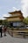 A traditional Japanese gold-colored pavilion surrounded by a serene garden landscape with trees and a gravel path. Two people are standing in conversation near a wooden fence in the foreground.
