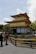 A traditional Japanese gold-colored pavilion surrounded by a serene garden landscape with trees and a gravel path. Two people are standing in conversation near a wooden fence in the foreground.