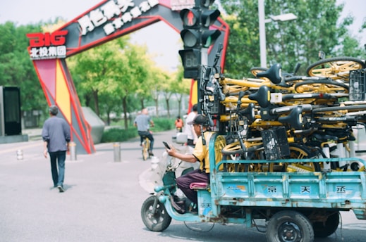 A small vehicle is piled high with rental bicycles, which have a yellow and black color scheme. The vehicle is traveling on a city street with a colorful archway in the background and a few people walking and riding bikes nearby.