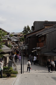 A bustling street lined with traditional wooden buildings, populated by tourists and locals walking and exploring the area. Some people are engaged in conversation, while others are capturing photos. The architecture and atmosphere suggest a historical district, with greenery visible on the sides.