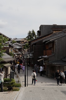 A bustling street lined with traditional wooden buildings, populated by tourists and locals walking and exploring the area. Some people are engaged in conversation, while others are capturing photos. The architecture and atmosphere suggest a historical district, with greenery visible on the sides.