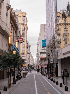 A bustling city street with people walking and old buildings lining both sides. The architecture features both classic and modern styles with a blend of ornate and reflective surfaces. Various shops and cafes are visible, along with signage in Spanish.