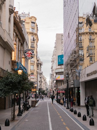 A bustling city street with people walking and old buildings lining both sides. The architecture features both classic and modern styles with a blend of ornate and reflective surfaces. Various shops and cafes are visible, along with signage in Spanish.