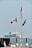 A cruise ship is visible in the distance, anchored near a rocky breakwater. In the foreground, a flagpole displays three flags: a United States flag at the top, a Canadian flag to the left, and an indiscernible blue flag to the right. Below, the dock is decorated with planters filled with pink and purple flowers.