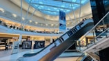 A modern shopping mall interior featuring a glass roof with structural beams and bright lighting. There are several retail stores visible, including Zara, and a prominent escalator descending across the frame. Shoppers are seen walking around and using the escalator. Large advertisements and banners are displayed throughout the space.