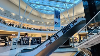 A modern shopping mall interior featuring a glass roof with structural beams and bright lighting. There are several retail stores visible, including Zara, and a prominent escalator descending across the frame. Shoppers are seen walking around and using the escalator. Large advertisements and banners are displayed throughout the space.