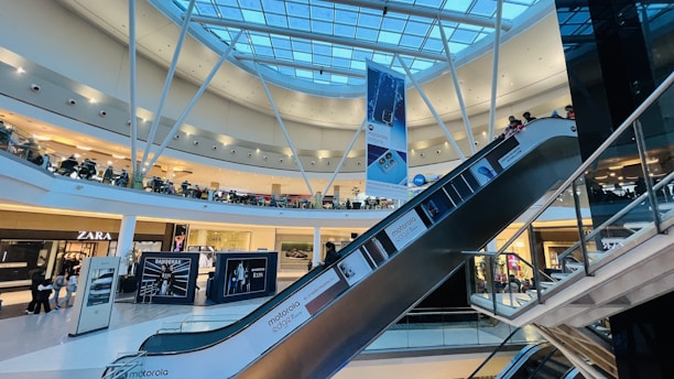 A modern shopping mall interior featuring a glass roof with structural beams and bright lighting. There are several retail stores visible, including Zara, and a prominent escalator descending across the frame. Shoppers are seen walking around and using the escalator. Large advertisements and banners are displayed throughout the space.