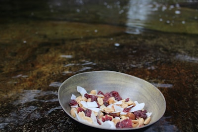 A bowl filled with a mix of dried fruits and nuts is placed on a wet surface. The bowl contains visible coconut flakes, almonds, and dried berries, against a blurred, water-textured background.
