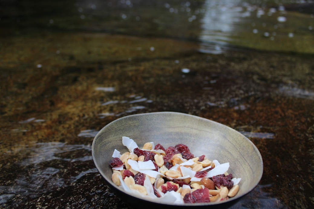 A bowl filled with a mix of dried fruits and nuts is placed on a wet surface. The bowl contains visible coconut flakes, almonds, and dried berries, against a blurred, water-textured background.
