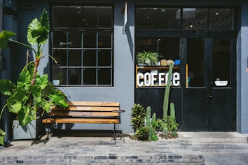 A quaint exterior of a coffee shop featuring a gray facade with a large window and black door. The word 'COFFEE' is displayed prominently in white letters on a shelf with potted plants inside the glass. A wooden bench sits outside next to a large tropical plant and a small side table, while several cacti and other plants are positioned near the entrance.