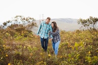 A couple laughing together while walking through a meadow filled with colorful flowers under a bright sky.