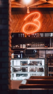 A cozy café interior featuring a glass display case filled with chocolate desserts, illuminated under soft warm lighting. Behind the counter is a wooden wall with a neon sign and various beverages and glassware arranged on shelves. A microwave and some other equipment are also visible.