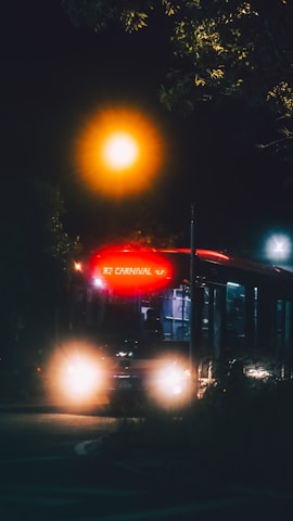 A bus is illuminated against the night sky, with its headlights on and a bright, glowing sign displaying '82 Carnival'. The surrounding area is dark, with a streetlight casting a warm, orange glow.