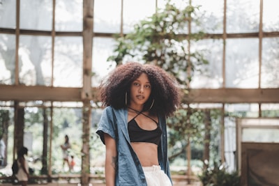 A woman with curly hair stands in a spacious indoor area with large windows, allowing natural light to flood in. She wears a blue denim shirt over a black top and white pants. Plants are visible in the background, contributing to a serene and natural ambiance.