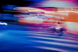 An abstract, blurred motion scene dominated by vibrant colors, including shades of blue, purple, and red. Light trails create dynamic patterns across the image, resembling a cityscape at night with streaks of light from vehicles and streetlights.