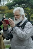 An elderly man with a white beard is capturing a moment with his smartphone outdoors. He is wearing a light grey jacket and has a DSLR camera hanging around his neck. The background features trees and greenery, suggesting a park or nature setting. Another person in the background also holds a smartphone, partially visible.
