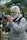 An elderly man with a white beard is capturing a moment with his smartphone outdoors. He is wearing a light grey jacket and has a DSLR camera hanging around his neck. The background features trees and greenery, suggesting a park or nature setting. Another person in the background also holds a smartphone, partially visible.